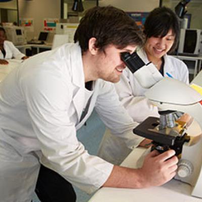 two students, dressed in lab coats, analysing a sample through a microscope.