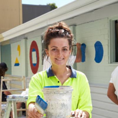 Painter with a pot of blue paint, standing in front of a wall where other painters are doing a mural