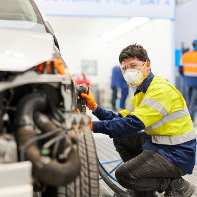 Student crouched down next to a car learning mechanics. Student is wearing safety goggles and mask