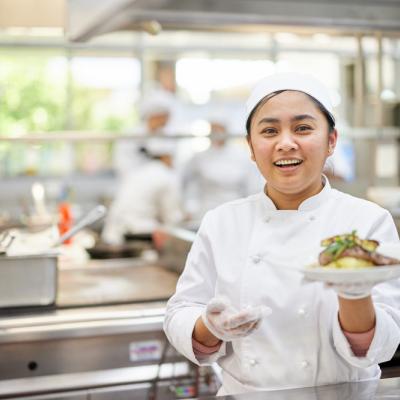 Cookery student dressed in white chef uniform holding up plate of food 