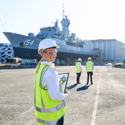 A student in maritime defence infront of ship wearing hard hat, safety goggles and high visibility shirt