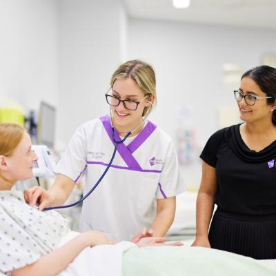 A nursing student checks a patient with a stethoscope, and another student stands beside her